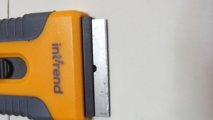 Ein Reinigungsschaber liegt auf einer glatten, hellen Oberfl&auml;che. Er wird verwendet, um K&uuml;chenkacheln hinter dem Herd zu s&auml;ubern.