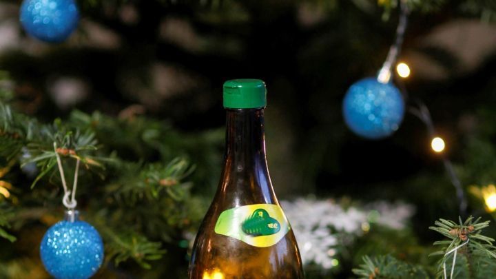 Eine Flasche Apfelessig steht vor einem festlich geschm&uuml;ckten Weihnachtsbaum mit blauen Kugeln und Lichtern.