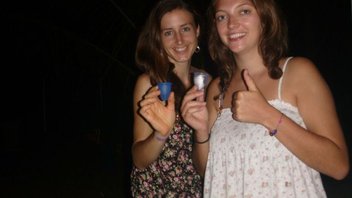 Zwei Personen halten Menstruationstassen in der Hand und l&auml;cheln in die Kamera im Dunkeln; eine zeigt den Daumen hoch.