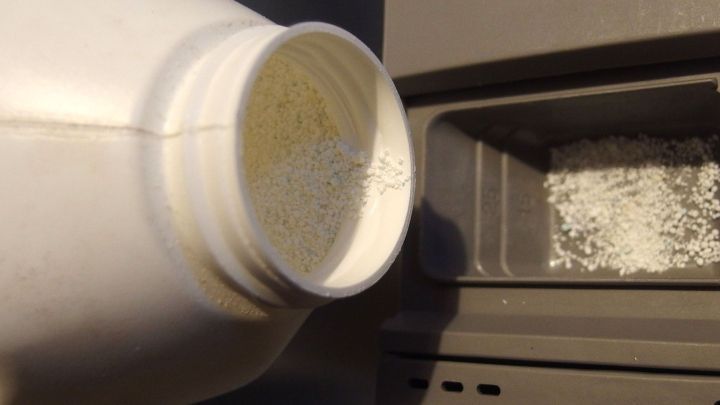 Ein wei&szlig;er Beh&auml;lter mit Geschirrsp&uuml;ler-Reiniger wird &uuml;ber das Fach eines Geschirrsp&uuml;lers gekippt, um die Vorrichtung zu bef&uuml;llen.