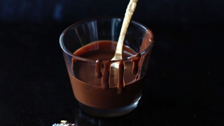 In einem Glas befindet sich geschmolzene Kuvert&uuml;re, w&auml;hrend ein L&ouml;ffel sie umr&uuml;hrt; Kekse liegen daneben.