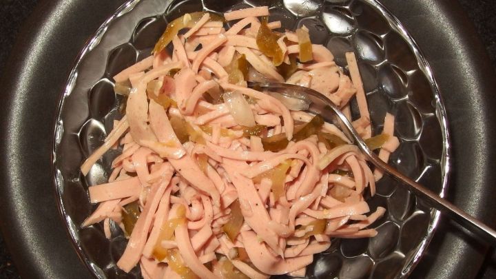 Wurstsalat liegt appetitlich in einer Sch&uuml;ssel, w&auml;hrend ein L&ouml;ffel die Mischung aus Wurst und Gurken umr&uuml;hrt.