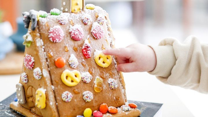 Ein kleines Kind ber&uuml;hrt eine bunt dekorierte Lebkuchenhaus-Dekoration mit vielen Bonbons in einer hellen Umgebung.