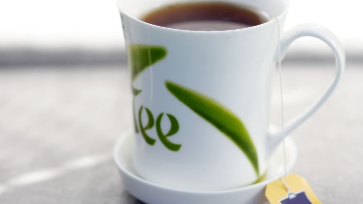 Eine Tasse mit schwarzem Tee steht auf einer Untertasse, mit einem Teebeutel, auf einer hellen Oberfl&auml;che.