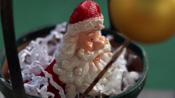 Ein Weihnachtsmann sitzt in einem kleinen Eimer, umgeben von wei&szlig;em Dekorationsmaterial, w&auml;hrend eine goldene Kugel schwingt.