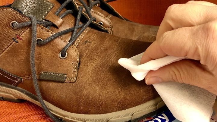 Ein Hand mit einem Feuchttuch reinigt den Schneerand eines braunen Schuhs auf einem orangefarbenen Hintergrund.