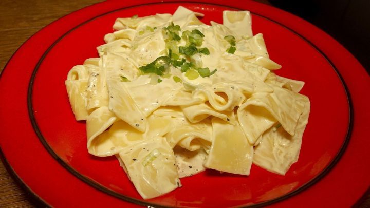 Eiernudeln sind auf einem roten Teller serviert, &uuml;berzogen mit einer cremigen K&auml;sesauce und garniert mit frischen Kr&auml;utern.