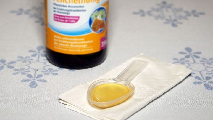 Ein Glas Fenchelhonig steht auf einem Tischtuch, w&auml;hrend ein L&ouml;ffel mit goldenem Inhalt daneben liegt.