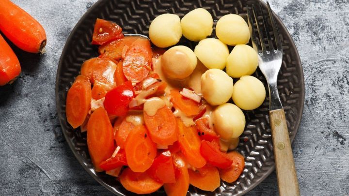 Auf einem dunklen Teller liegt M&ouml;hrengem&uuml;se mit Tomaten und Kartoffelkn&ouml;deln, angerichtet auf einer grauen Oberfl&auml;che.
