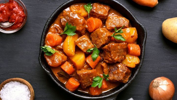 Ein k&ouml;stlicher M&ouml;hreneintopf mit Rindfleisch und frischem Gem&uuml;se wird in einer gusseisernen Pfanne serviert, umgeben von Kartoffeln und Zutaten.