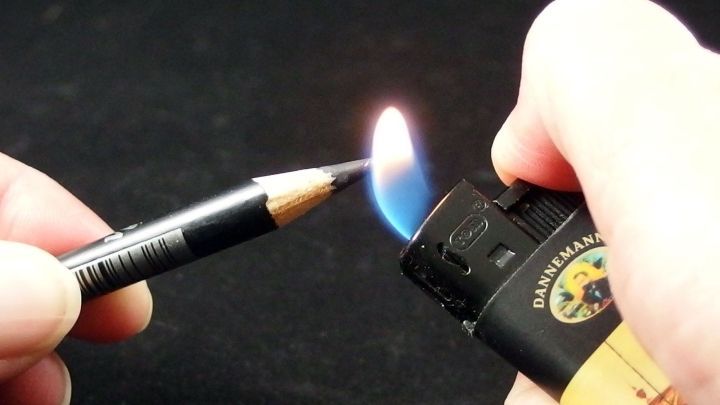 Ein Feuerzeug entz&uuml;ndet die Spitze eines Kajalstifts, w&auml;hrend eine Hand ihn h&auml;lt, auf einem schwarzen Hintergrund.