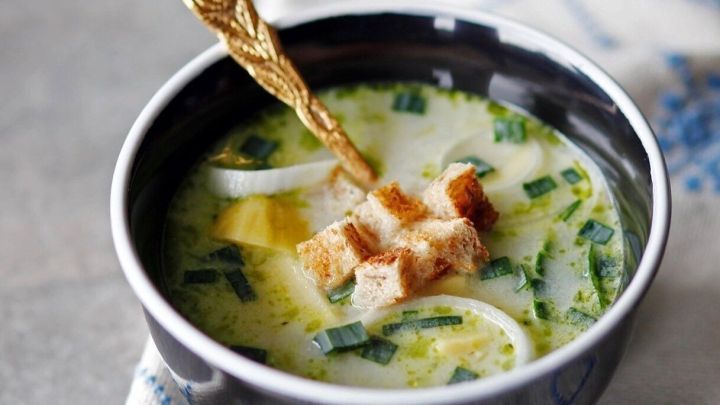 Eine cremige Kartoffel-Lauchsuppe in einer schwarzen Schale, garniert mit Brotw&uuml;rfeln und einem goldenen L&ouml;ffel.