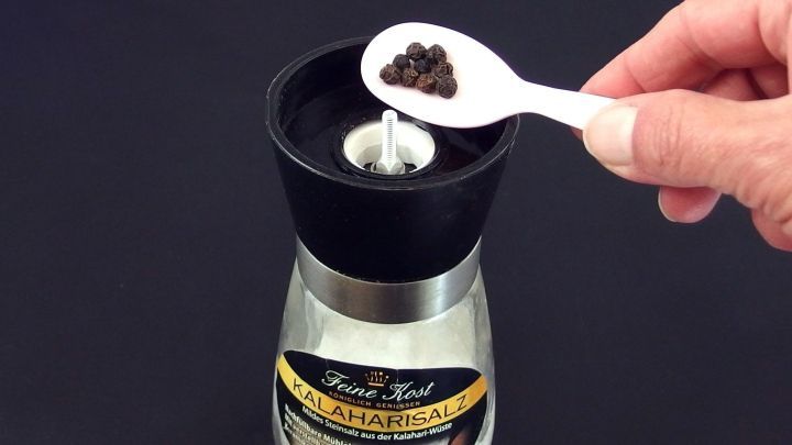 Eine Hand f&uuml;gt mit einem L&ouml;ffel Pfefferk&ouml;rner in eine M&uuml;hle, die bereit ist, um Salz und Pfeffer zu mahlen.