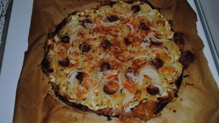 Eine Pizza Teutonica liegt auf Backpapier, reich belegt mit Wurst, Zwiebeln und K&auml;se, in einer hellen K&uuml;che.