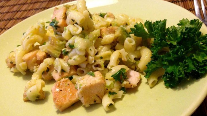 Ein Nudelsalat mit gebratenem Lachs und Avocado liegt auf einem hellgr&uuml;nen Teller, umgeben von frischer Petersilie.