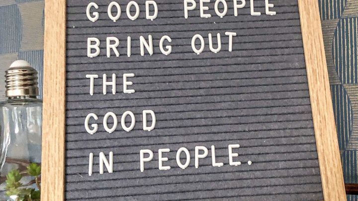 Ein Holzschild steht auf einem Tisch und zeigt den Text &bdquo;GOOD PEOPLE BRING OUT THE GOOD IN PEOPLE.&ldquo; in einer gem&uuml;tlichen Umgebung.