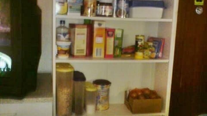 Ein schmaler Regal mit Lebensmitteln und Kochutensilien steht in einer kleinen K&uuml;che, w&auml;hrend ein Fernseher im Hintergrund l&auml;uft.