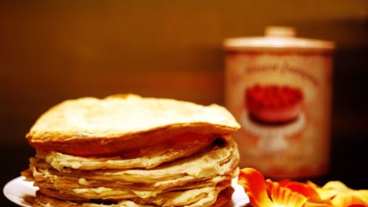 Eine kalte Pfannkuchentorte mit R&auml;ucherlachs liegt auf einem Teller, umgeben von bunten Bl&uuml;tenbl&auml;ttern und einer Dose im Hintergrund.