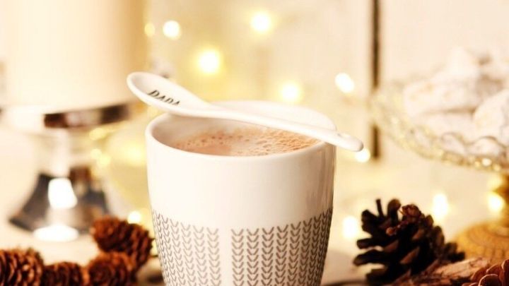 Eine Tasse Winterkakao steht auf einem festlichen Tisch, umgeben von Teelichtern, Tannenzapfen und Zimtstangen.