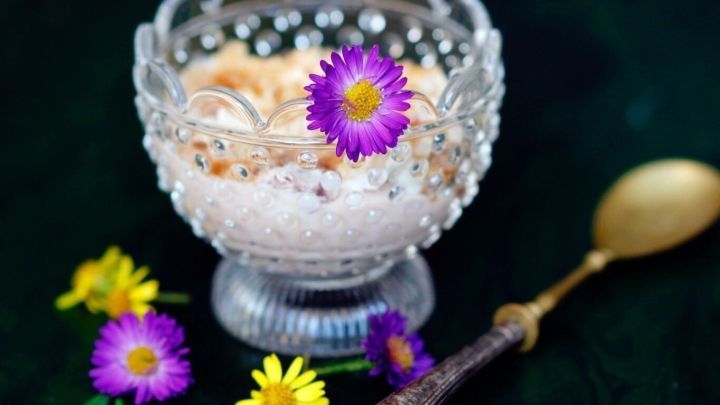 Eine elegante Glasschale enth&auml;lt eine sahnige Nachspeise, die mit bunten Blumen dekoriert ist, auf einem dunklen Untergrund.