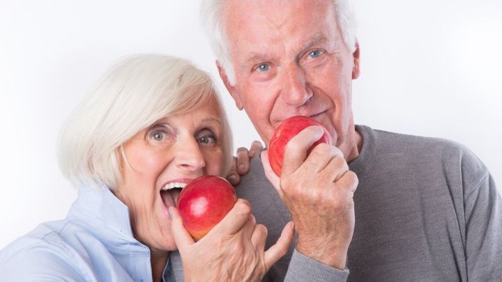 Ein &auml;lteres Paar h&auml;lt jeweils einen roten Apfel und bei&szlig;t hinein, l&auml;chelnd vor einem neutralen Hintergrund.