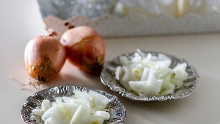 H&auml;ufchen gehackter Zwiebeln liegen auf silbernen Tellern, w&auml;hrend frische Zwiebeln im Hintergrund sichtbar sind.