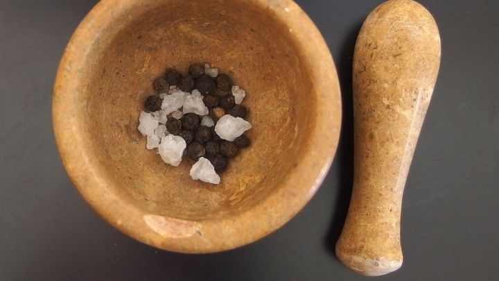 Ein Holz-M&ouml;rser enth&auml;lt schwarze Pfefferk&ouml;rner und grobes Salz, w&auml;hrend der St&ouml;&szlig;el daneben liegt, bereit zum Zerkleinern.