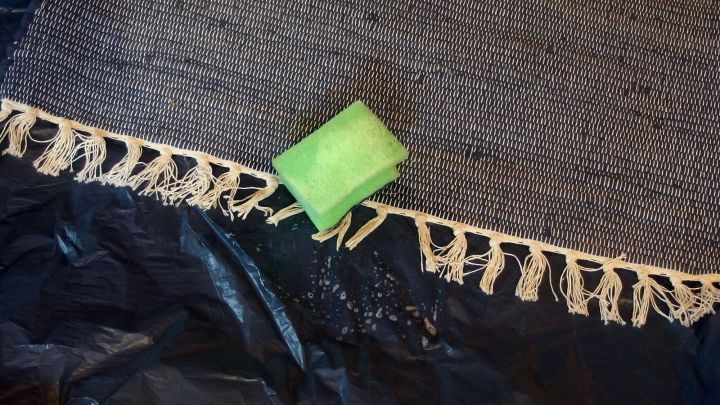 Ein Teppich liegt auf einem schwarzen Untergrund, w&auml;hrend ein gr&uuml;ner Schwamm &uuml;ber die Teppichfransen gereinigt wird.