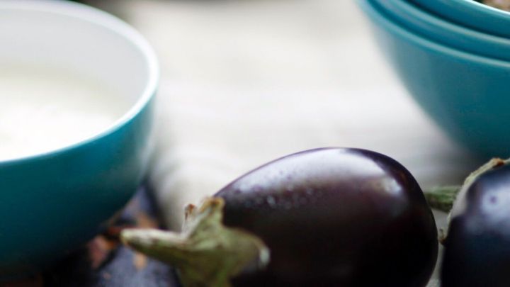 Eine gl&auml;nzende Aubergine liegt neben einer Sch&uuml;ssel mit einer gebrauchsfertigen Sauce, umgeben von weiteren Zutaten auf einem Holzbrett.