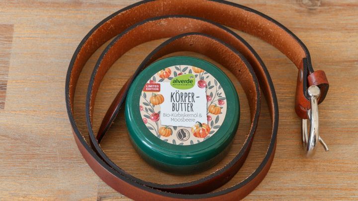 Eine gr&uuml;ne Dose mit der Aufschrift "K&ouml;rperbutter" liegt auf einem Holztisch, umgeben von einem braunen G&uuml;rtel.