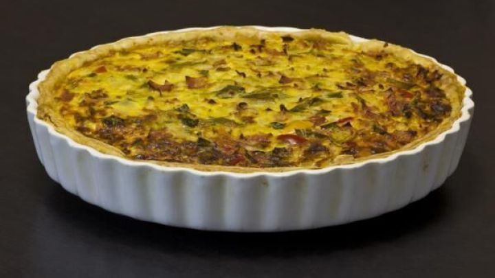 Eine goldbraun gebackene Quiche Lorraine ruht in einer wei&szlig;en Tarteform auf einer dunklen Oberfl&auml;che, bereit zum Servieren.