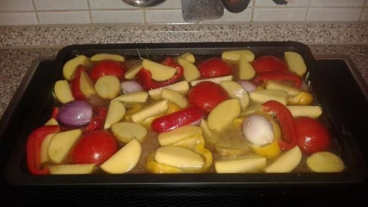 Bunte Kartoffel-Paprika-Gem&uuml;se-Mischung liegt auf einem Blech, umgeben von K&uuml;chenger&auml;ten und Holztisch.