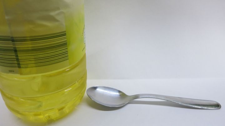 Eine Flasche Sonnenblumen&ouml;l steht neben einem silbernen L&ouml;ffel auf wei&szlig;em Hintergrund, bereit zur Nutzung.