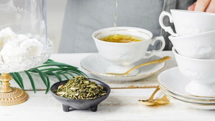 Eine Teekanne gie&szlig;t hei&szlig;e Kr&auml;uterteemischung in eine feine Tasse, umgeben von einem Teller mit getrockneten Kr&auml;utern.