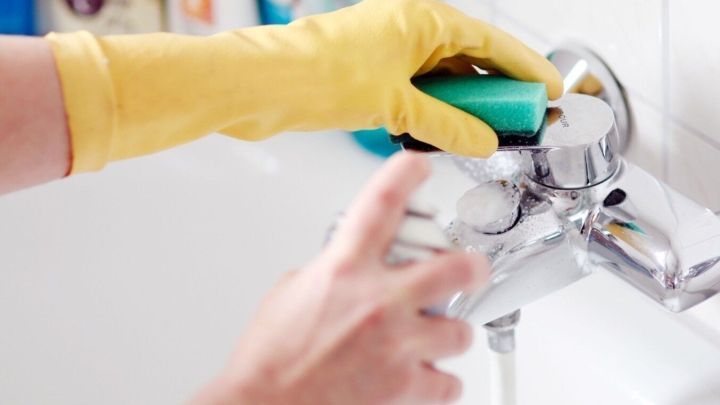 Eine Hand mit einem gelben Gummihandschuh reinigt einen Wasserhahn mit Backofenspray in einem hellen Badezimmer.