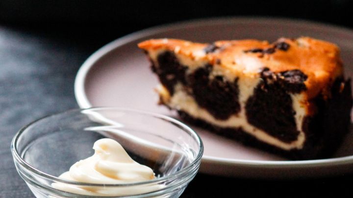 Ein saftiges St&uuml;ck Kuchenteig auf einem Teller liegt neben einer Sch&uuml;ssel mit Mayonnaise, auf einem dunklen Tisch.