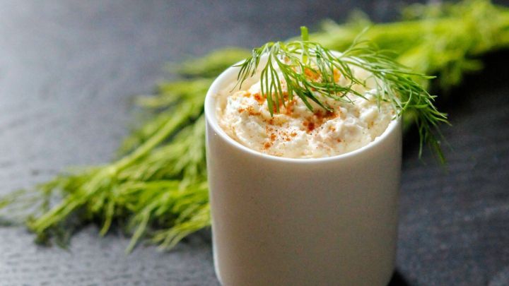 Ein ged&auml;mpfter Gem&uuml;se-Dip steht in einem wei&szlig;en Becher, garniert mit frischem Dill, auf einem dunklen Holzuntergrund.
