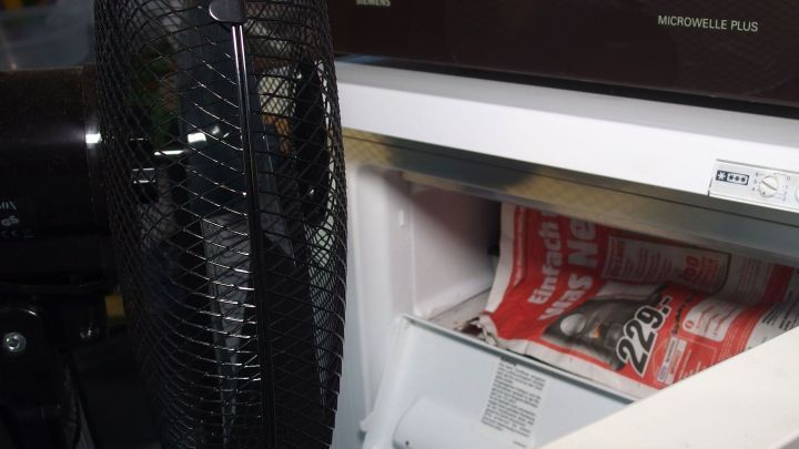 Ein Ventilator steht neben einem Gefrierschrank, der offen ist und eine Zeitschrift in der unteren Schublade zeigt.
