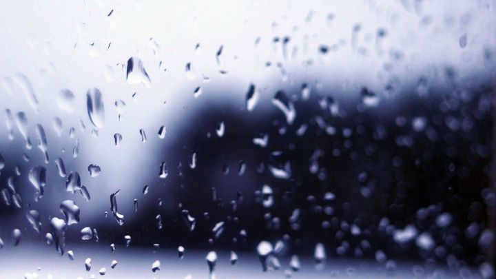 Wassertropfen perlen auf einem Fenster, wodurch die Sicht verschwommen wird und ein grauer, nebliger Hintergrund sichtbar ist.