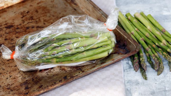 Gr&uuml;ner Spargel in einer Plastikt&uuml;te liegt auf einem Backblech, w&auml;hrend frischer Spargel daneben ausgebreitet ist.