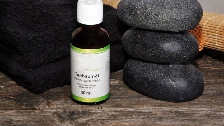 Eine Flasche Teebaum&ouml;l steht neben vier schwarzen Steinen auf einer Holzoberfl&auml;che, umgeben von einem schwarzen Handtuch.