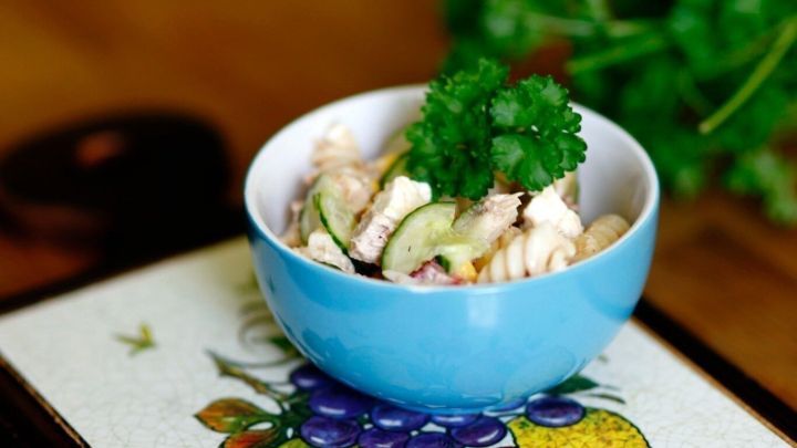 Ein blauer Sch&uuml;ssel enth&auml;lt Nudelsalat mit Gurken, garniert mit frischem Koriander, auf einem bunten Tischuntersetzer.