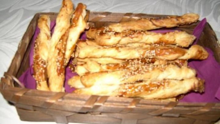 Sesamstangerl liegen in einem braunen Korb auf lila Serviette. Sie sind mit Salz bestreut und wirken knusprig.