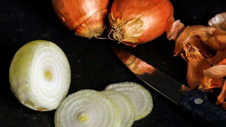 Eine geschnittene Zwiebel liegt neben ganzen Zwiebeln und einem Messer auf einer dunklen Arbeitsfl&auml;che, bereit zum R&ouml;sten.