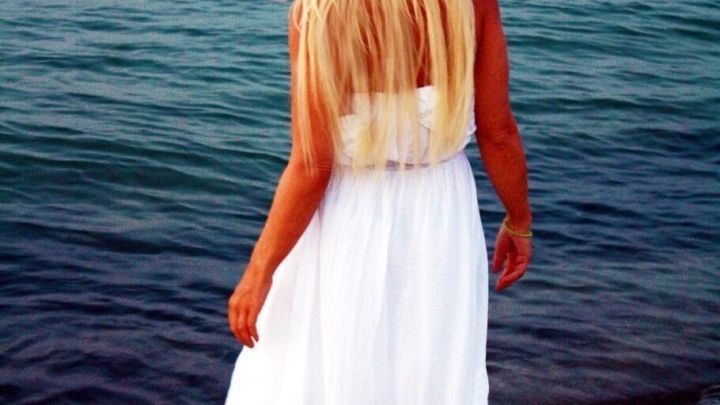 Eine blonde Frau im wei&szlig;en Kleid steht barfu&szlig; im flachen Wasser am Strand und genie&szlig;t den Sonnenuntergang.