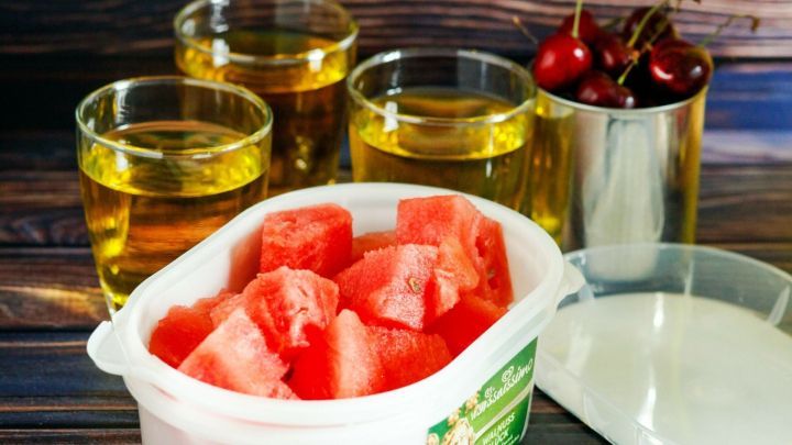 In einer Sch&uuml;ssel liegt frisches Wassermelonens St&uuml;ck, w&auml;hrend Gl&auml;ser mit Getr&auml;nken und Kirschen auf einem Holzuntergrund stehen.