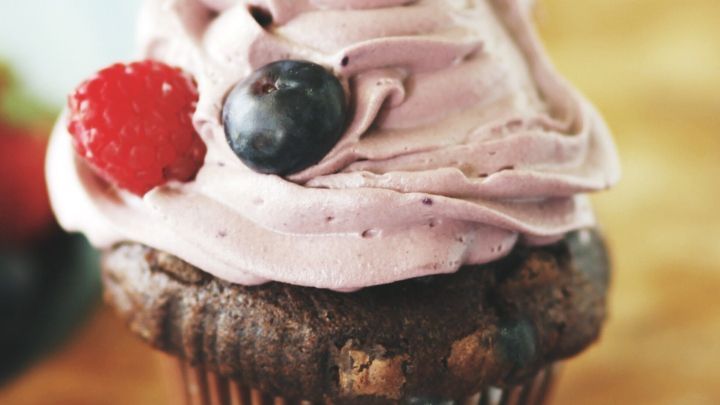 Ein saftiger Blaubeermuffin mit einem bunten, cremigen Frosting steht auf einem Holzbrett in heller Umgebung.
