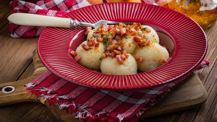 Kartoffelkl&ouml;&szlig;e liegen auf einem roten Teller, garniert mit Speck und Petersilie, umgeben von rustikalem Holz und dekorativen T&uuml;chern.