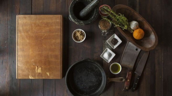 Ein M&ouml;rser aus Stein steht auf einem Holztisch, umgeben von Gew&uuml;rzen und frischen Zutaten f&uuml;r die Zubereitung.