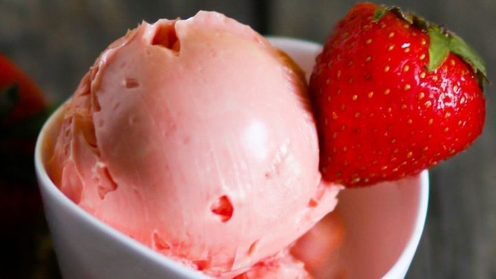 Eine Schale mit Erdbeereis, dekoriert mit einer frischen Erdbeere, steht auf einem rustikalen Holztisch.